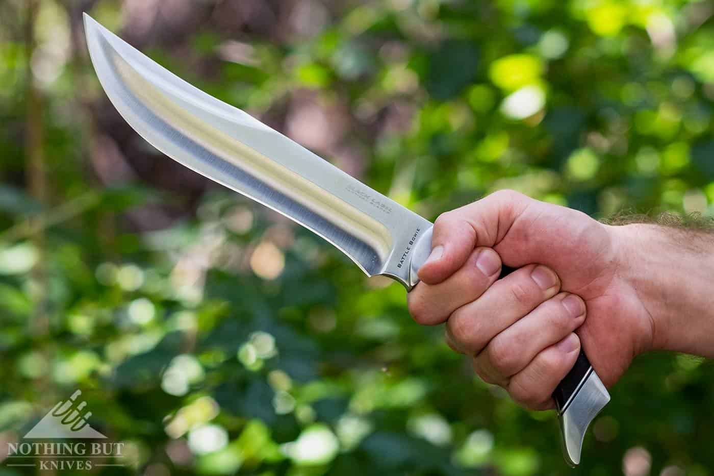 A close-up of a man's hand holding a Browning Battle Bowie knife with green foliage in the background. 