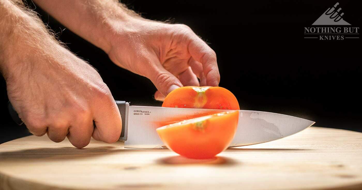 The Cold Steel Kitchen Classics 8 Inch Chef Knife slicing through a tomato on a wood cutting board in front of a black background.