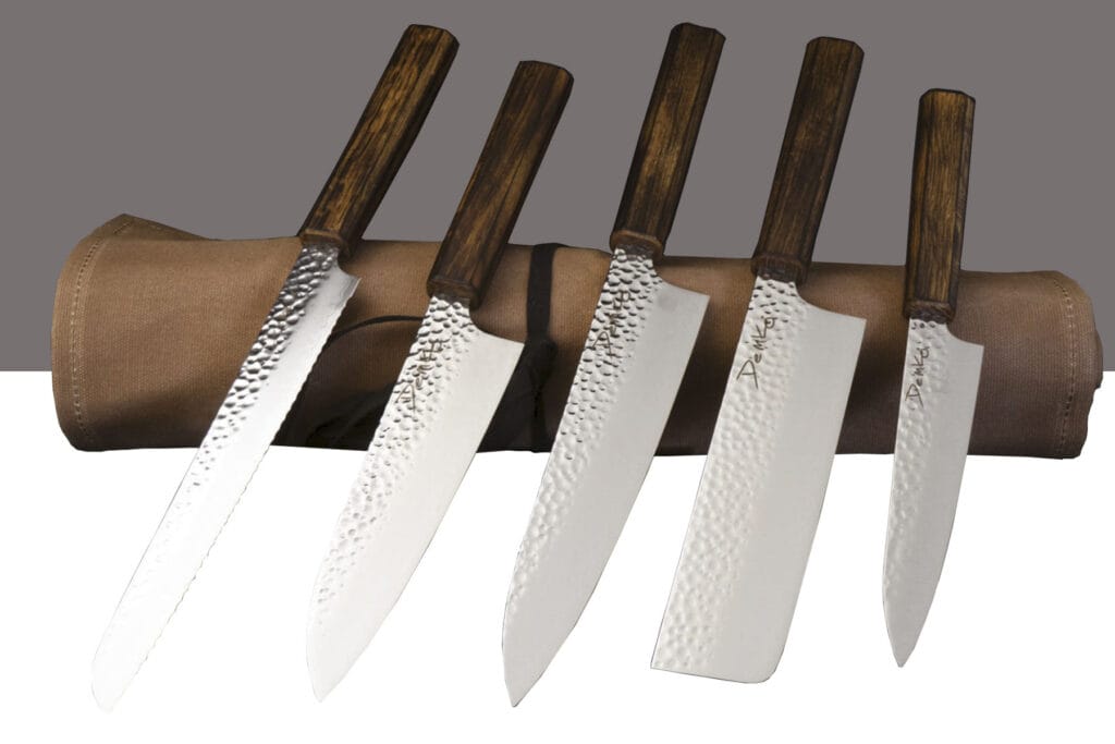 Our Ongoing Hunt For Great Japanese Knife Sets - Image 28 The fives knives of the Demko kitchen cutlery set shown here on top of their wax canvas storage roll in front of a two tone white and grey background.