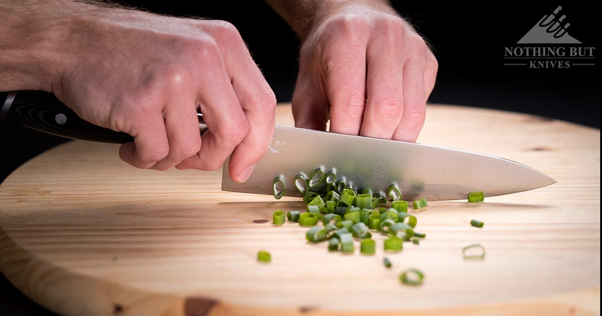 A man's hand shown dicing green onions on a wooden cutting board with the Yaxell Mon Chef knife.