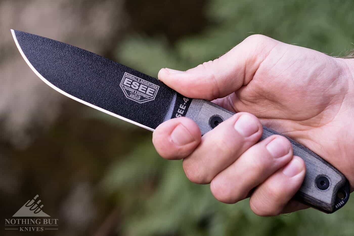 The Esee 4 knife with a 1095 high carbon steel blade in a man's hand outdoors. 