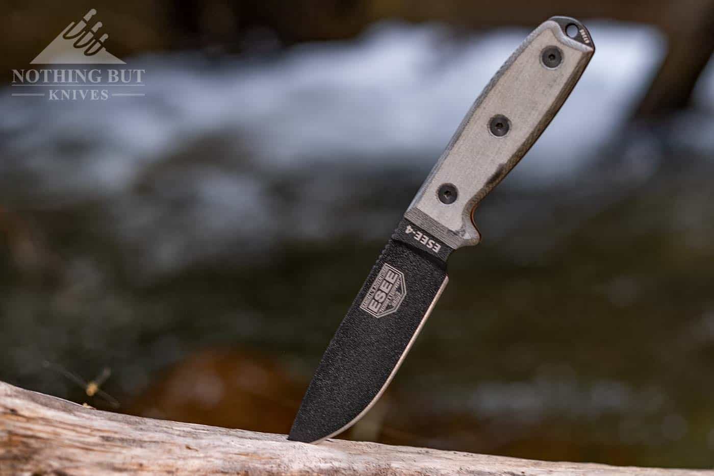 The Esee 4 tactical survival fixed blade knife sticking out of a log on the edge of a small mountain creek. 