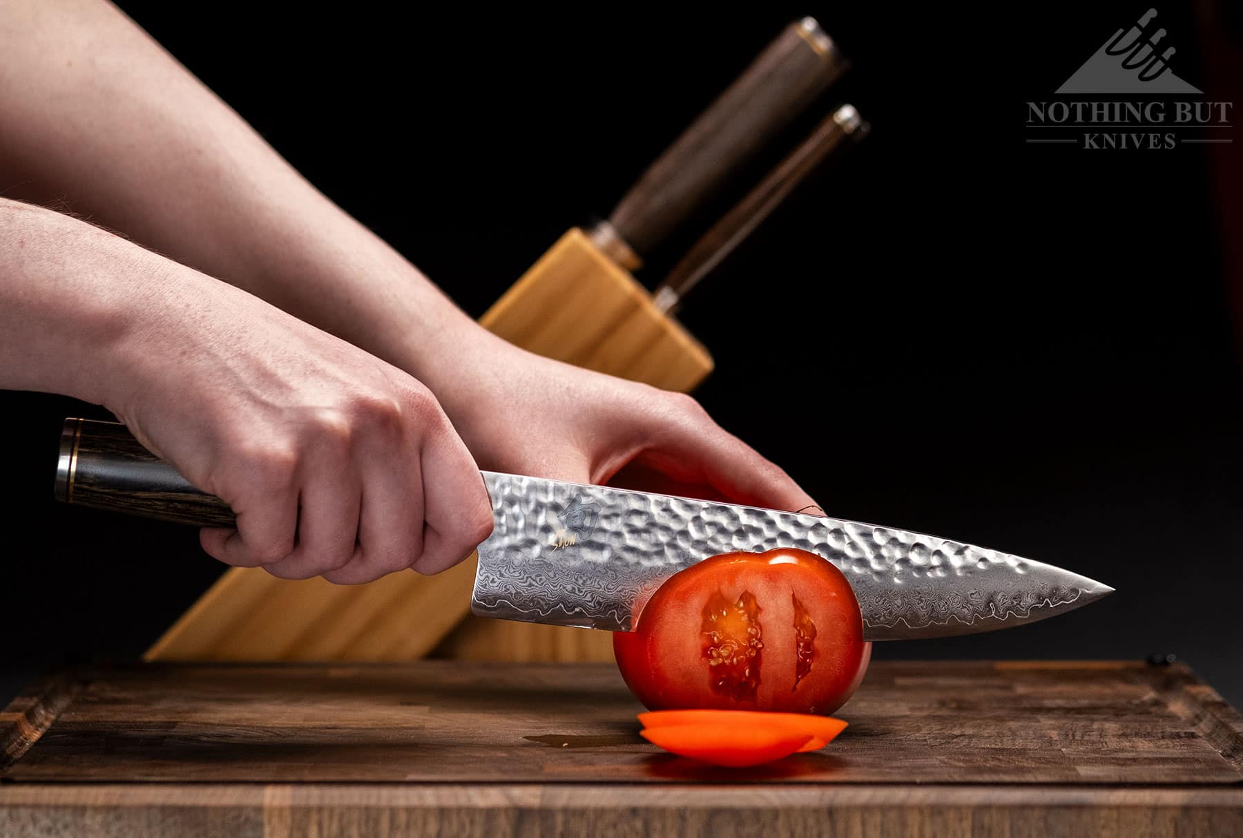A chef knife from the Shun Premier Japanese knife set being used to slice a tomato in front of the storage block.
