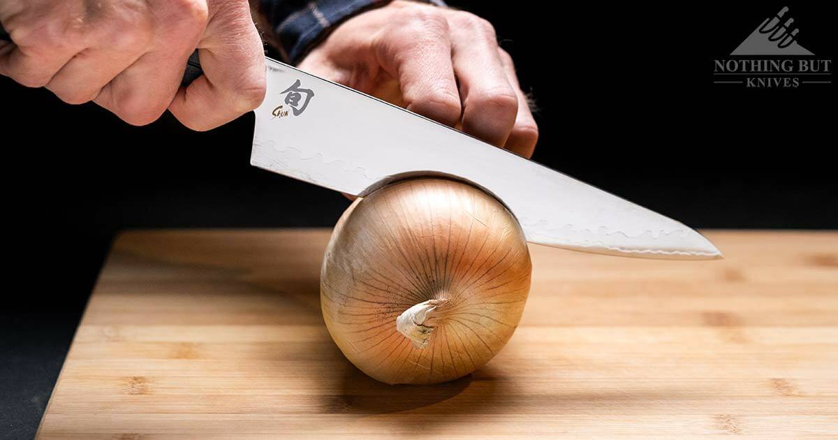 A close up of a man's hand using the Shun Sora chef knife to cut through a yellow onion. 