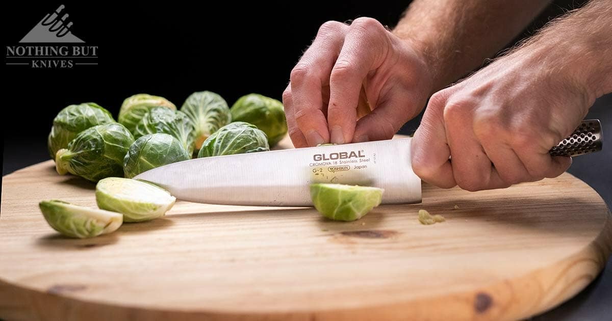 A close-up of a man's hand slixing Brissel sprouts with a Global chef knife. 