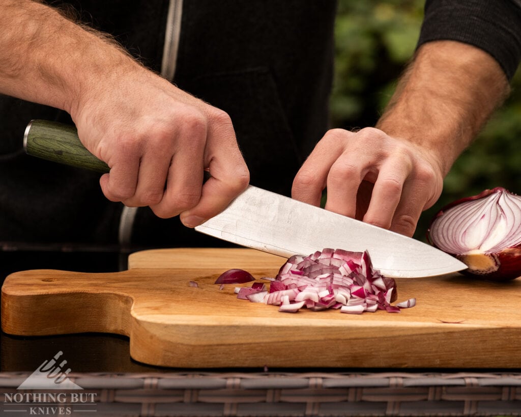 Best High-End Knife Sets For Professional Or Home Use - Image 12 A close-up of the 8-inch Hexclad chef knife being used to dice a red onion on a wood cutting board.