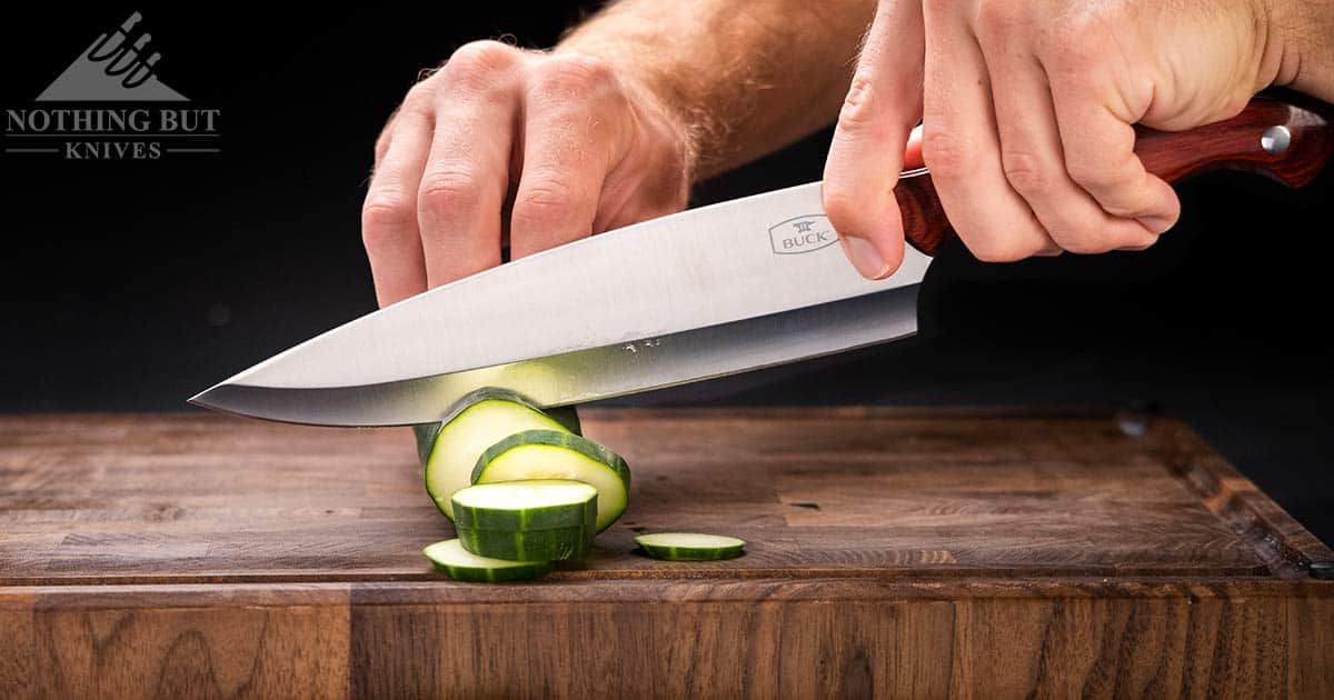 Best Professional And High End Kitchen Knife Sets In 2023 33 A close-up of the 8 inch Buck chef knife with rosewood handle slicing through a cucumber on a wood cutting board.