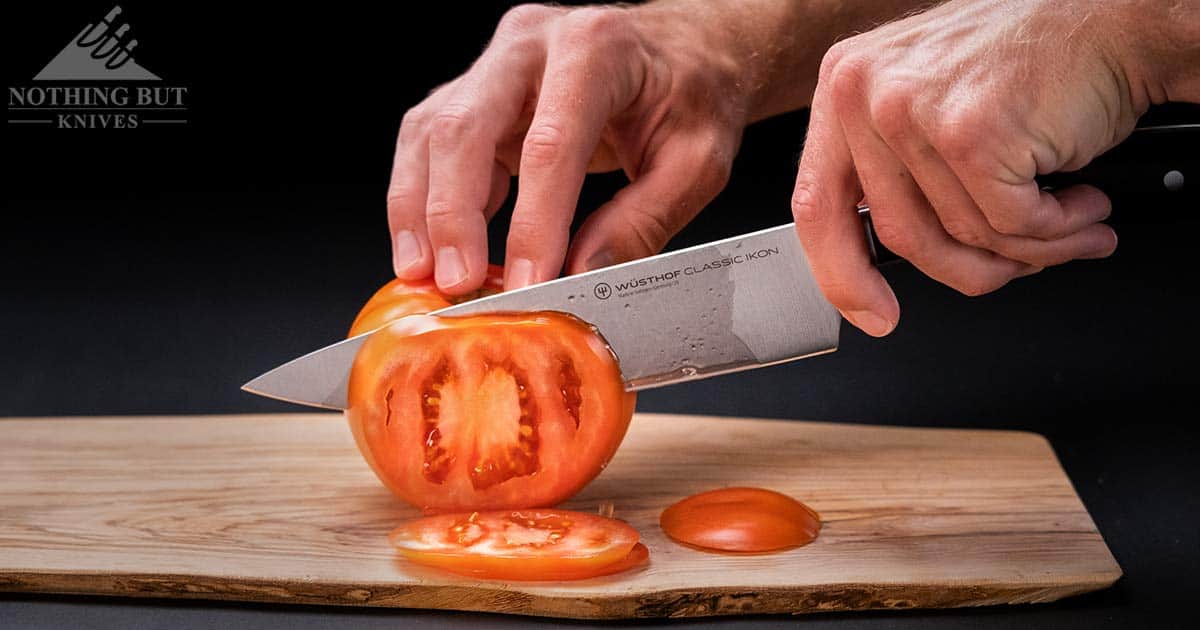 The Classic Ikon 8 inch chef knife slicing through a tomato on a wood cutting board in front of a dark background. 