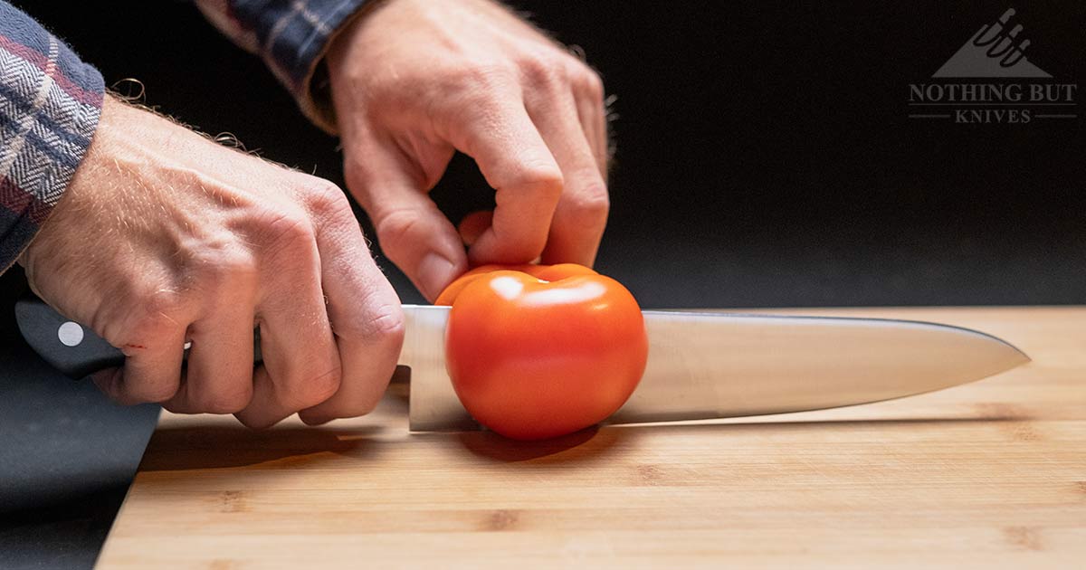A Man's hand holding a Tojiro chef knife that is salicing through a tomato on a cutting board.