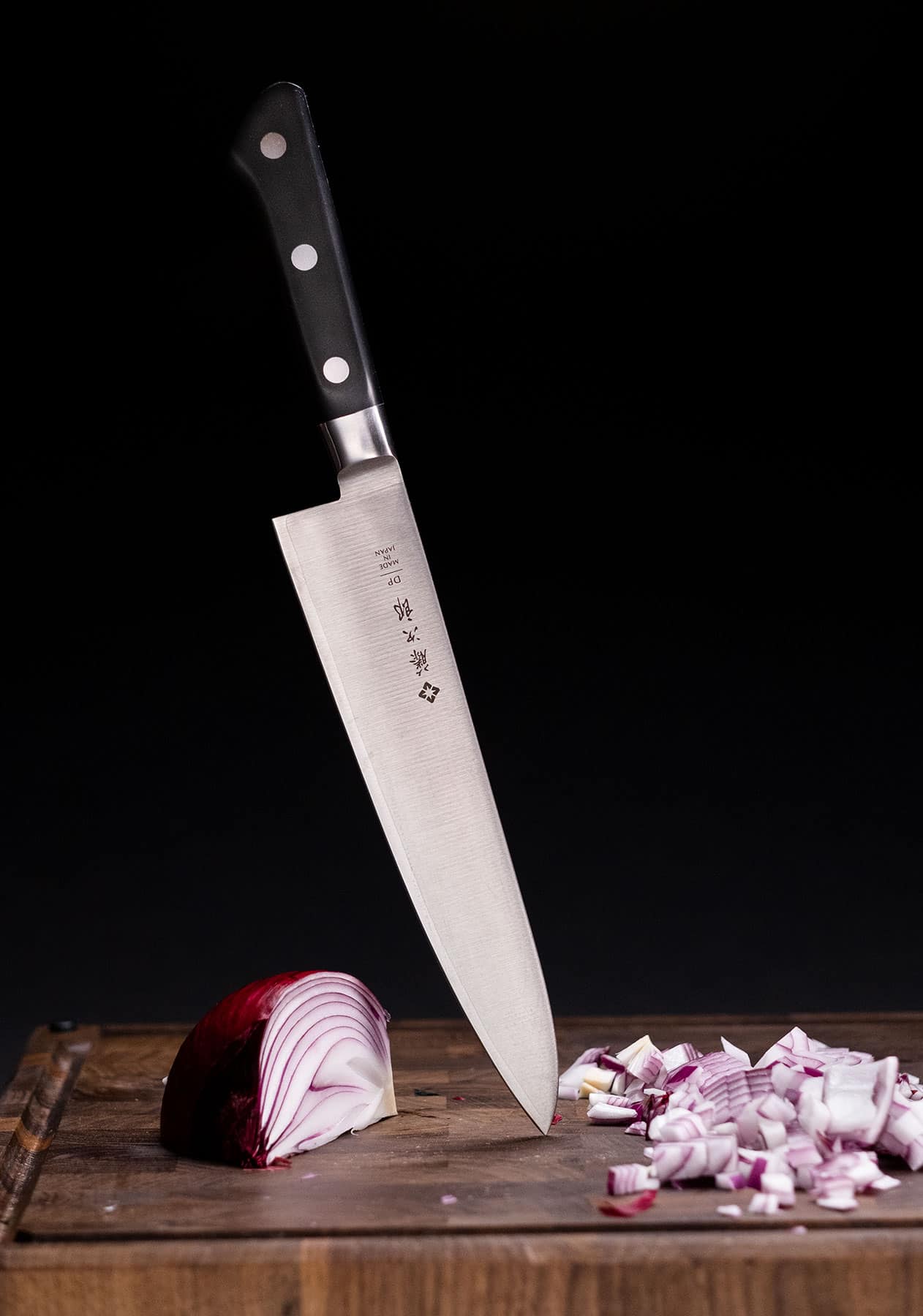 The Tojiro DP 8-inch chef standing on its blade tip on a dark wood cutting board with a red onion that has been diced in front of a black background.