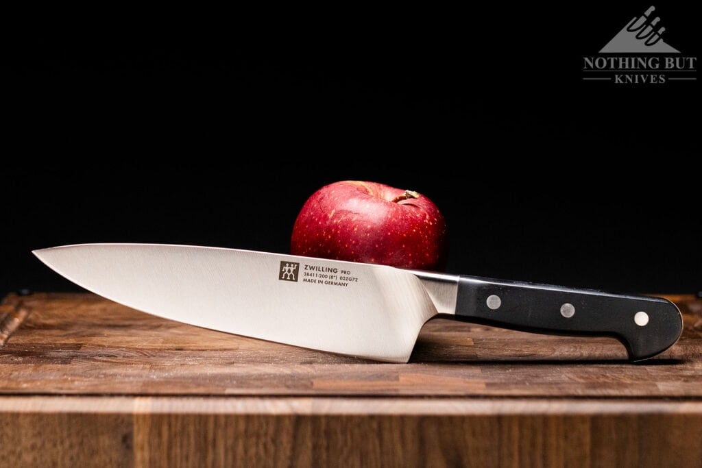 Best High-End Knife Sets For Professional Or Home Use - Image 9 The Zwilling Pro 8-inch chef knife with a red apple on a wood cutting board in front of a dark background.