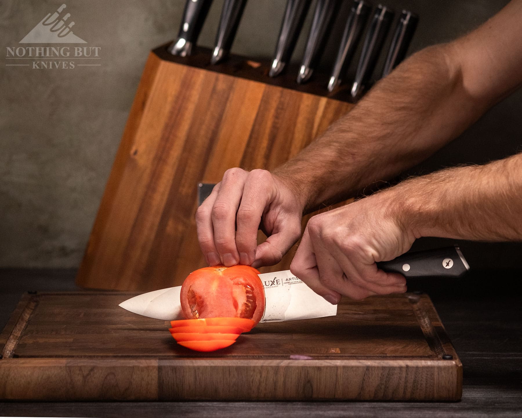 Best Kitchen Knife Sets Under $300 - Image 5 The Cutluxe Artisan 8-inch chef knife being used to slice a tomato on an end grain cutting board in front of the 8-piece knife set.