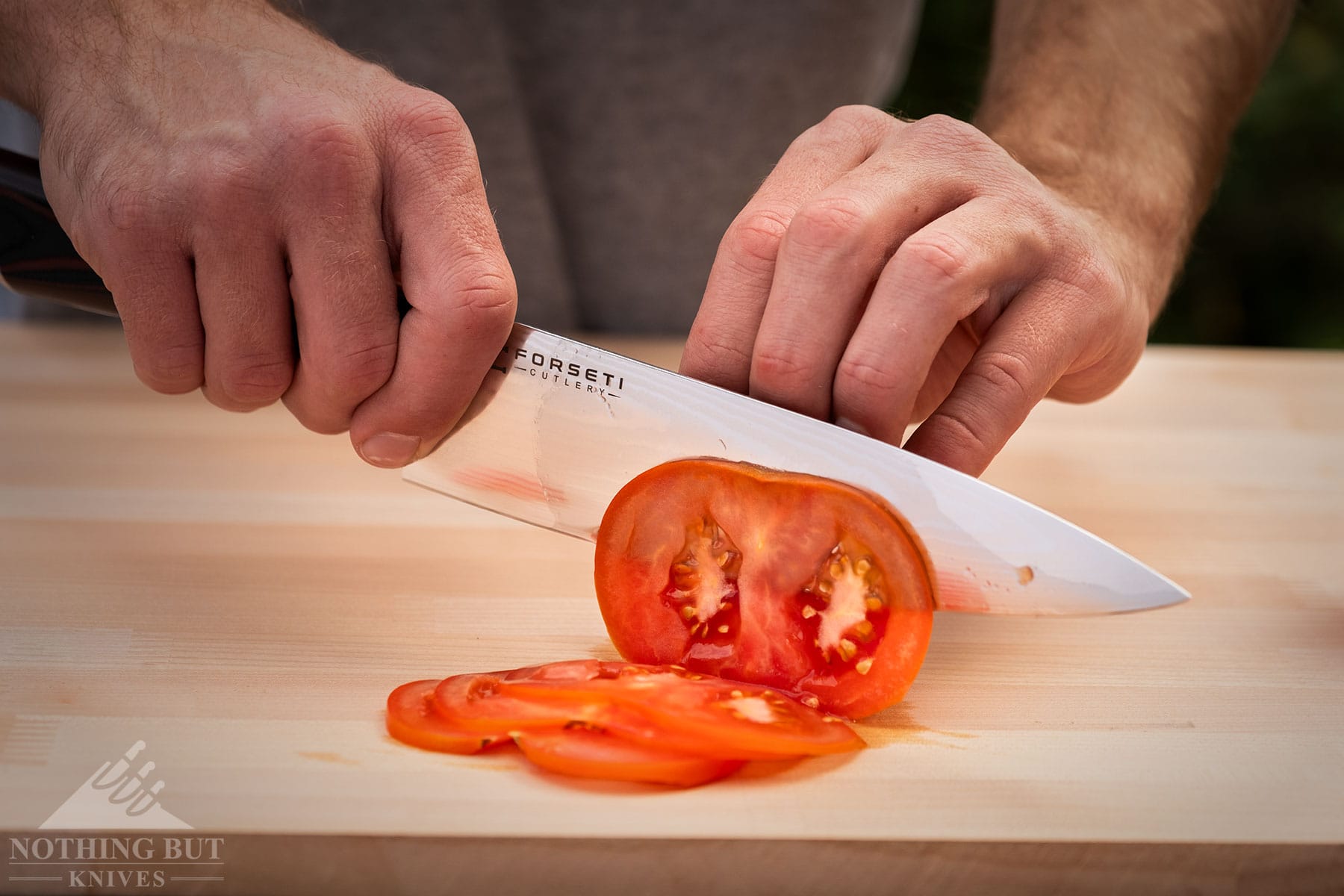 Best Kitchen Knife Sets Under $300 - Image 10 An elevated, side view of the Forsti Cutlery 8-inch chef knife being used to slice a tomato on a bamboo wood cutting board.