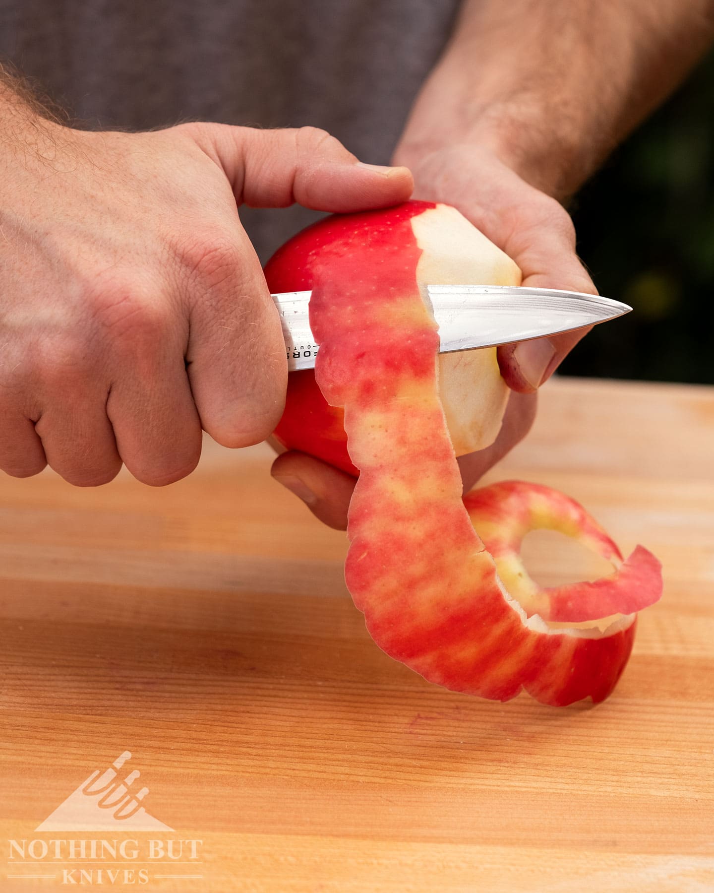 Best Kitchen Knife Sets Under $300 - Image 11 A close-up of the Forseti Cutlery paring knife being used to peel a red apple above a Bamboo wood cutting board.