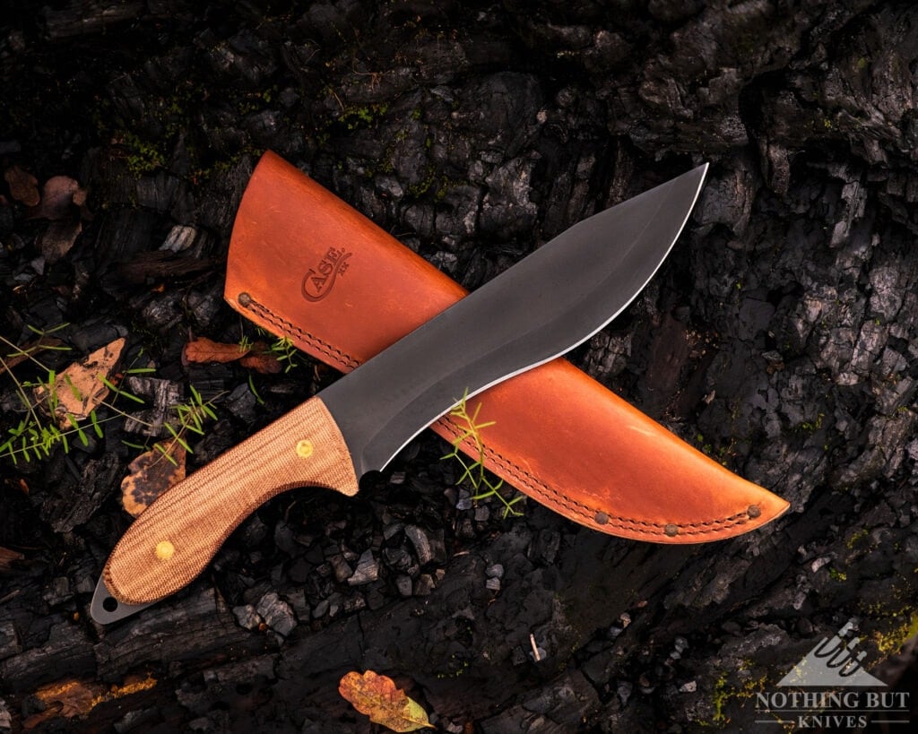 An overhead view of a large Bowie knife that was made in the USA shown here with its brown leather sheath on a charred log.