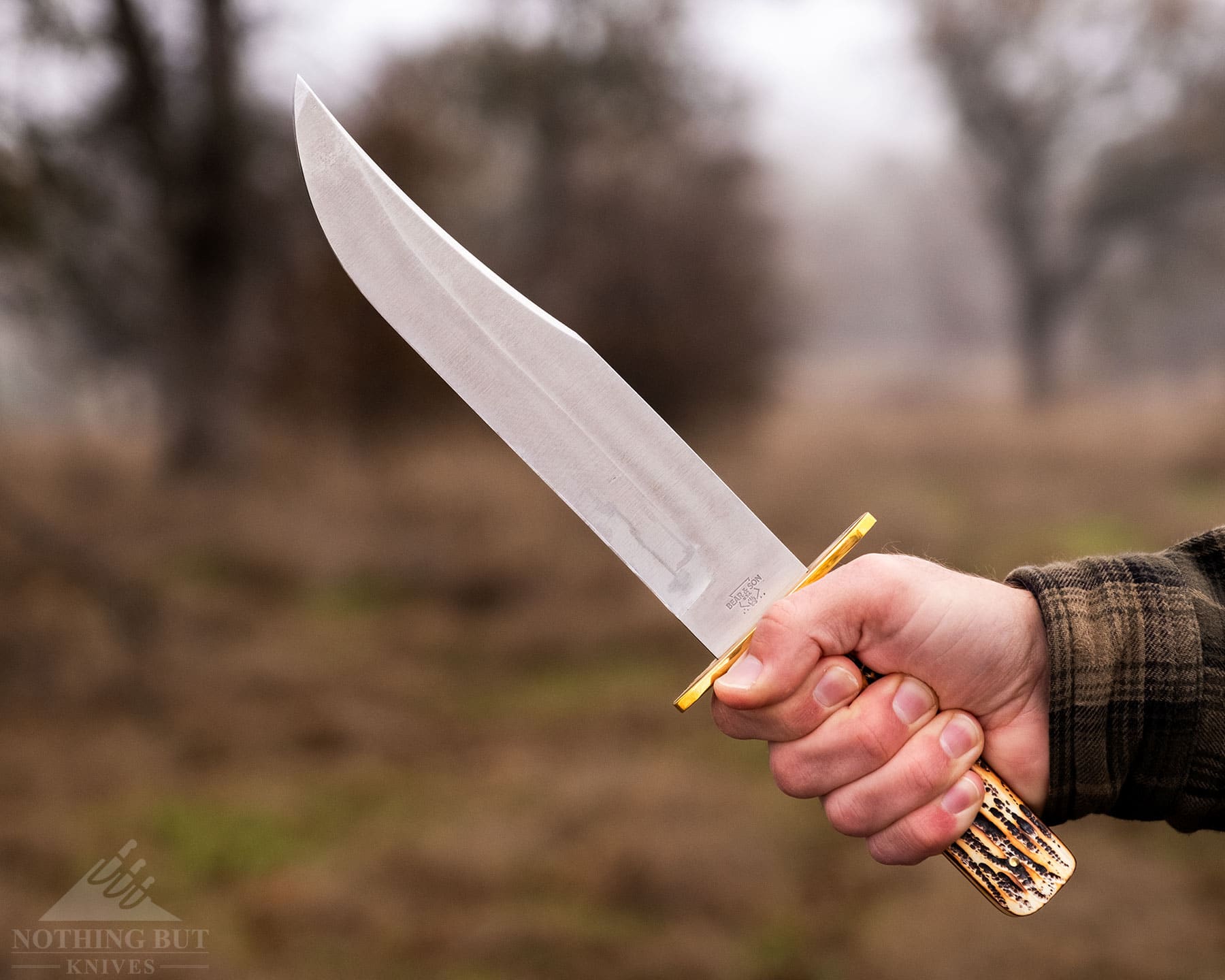 A close-up of the BEar & Son 9-inch Bowie in a person's right hand in front of a forest on an overcast day. 
