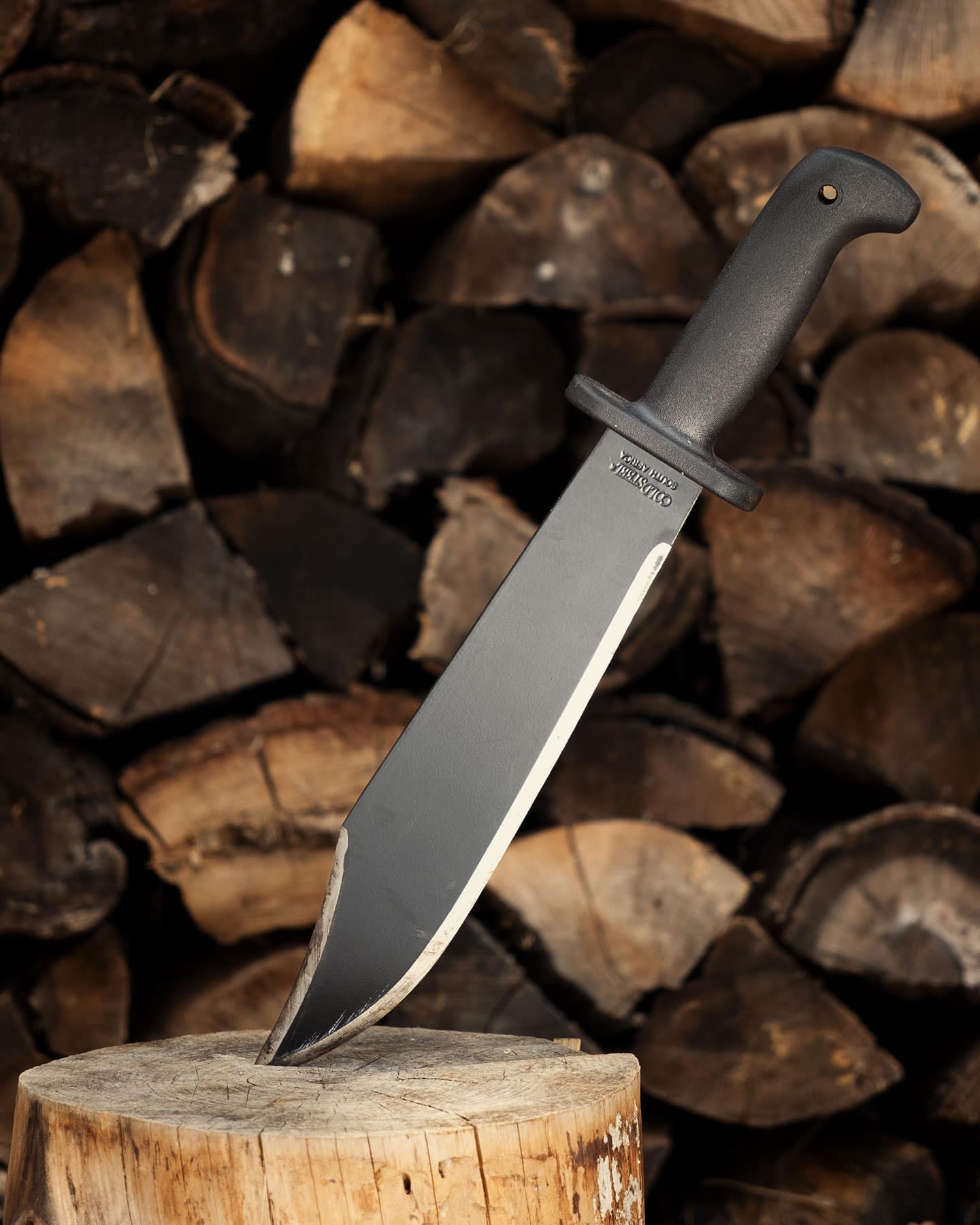The Cold Steel Black Bear Bowie knife with its tip stuck in a tree stump in front of a pile of firewood.