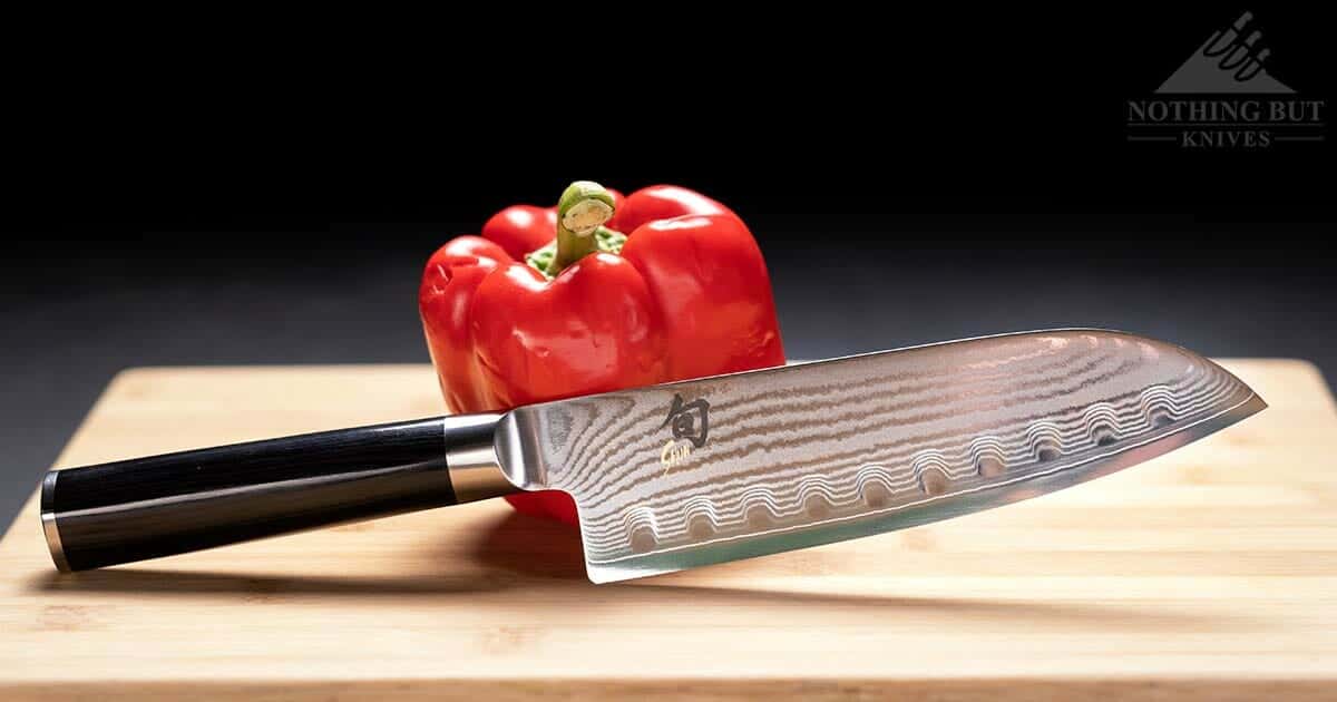 The Shun Classic Santoku Knife leaning againsts a red pepper on a wood cutting board.