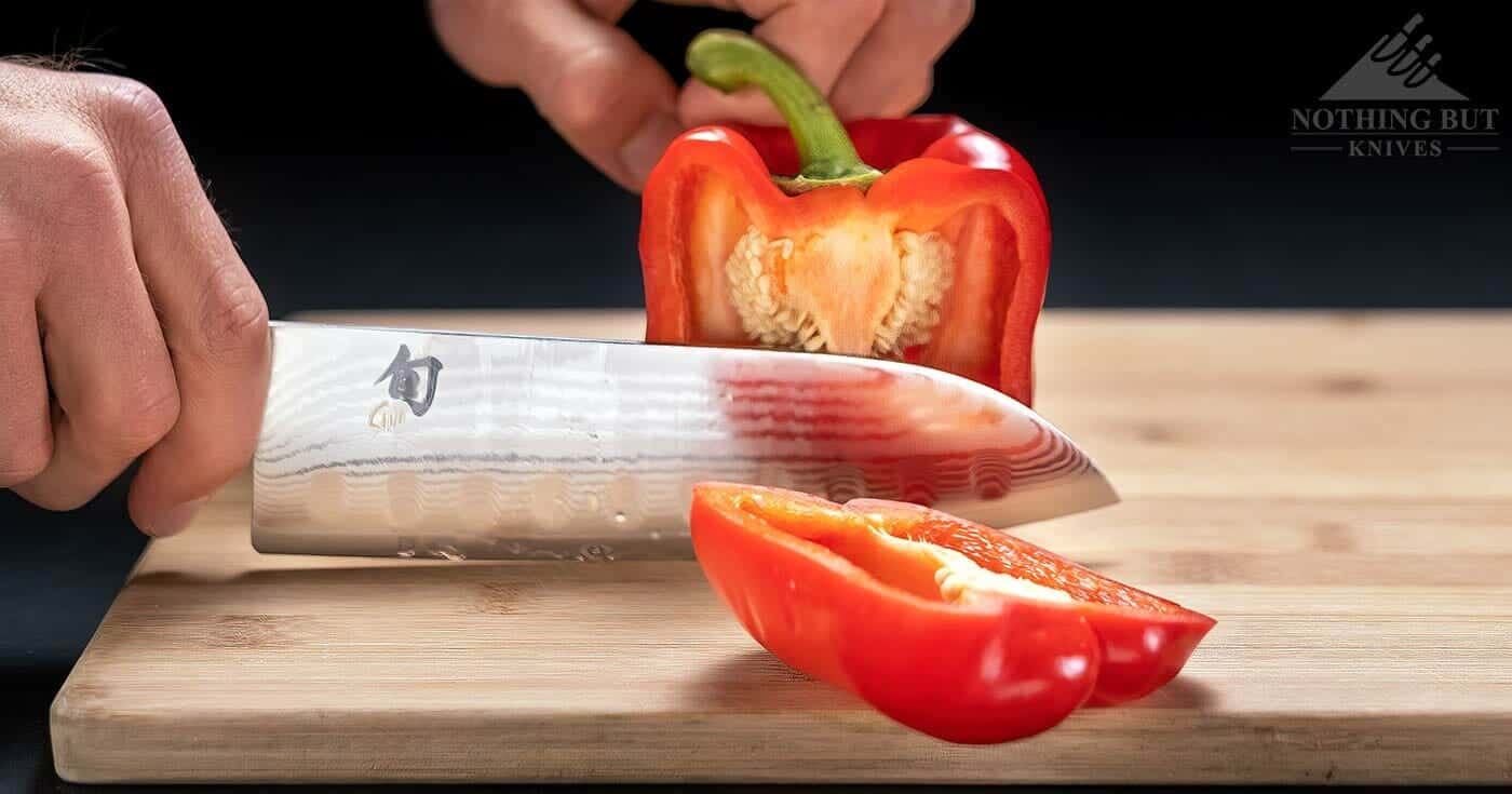 The Shun Classic Santoku knife alicing through a red pepper on a wood cutting board on a black background. 