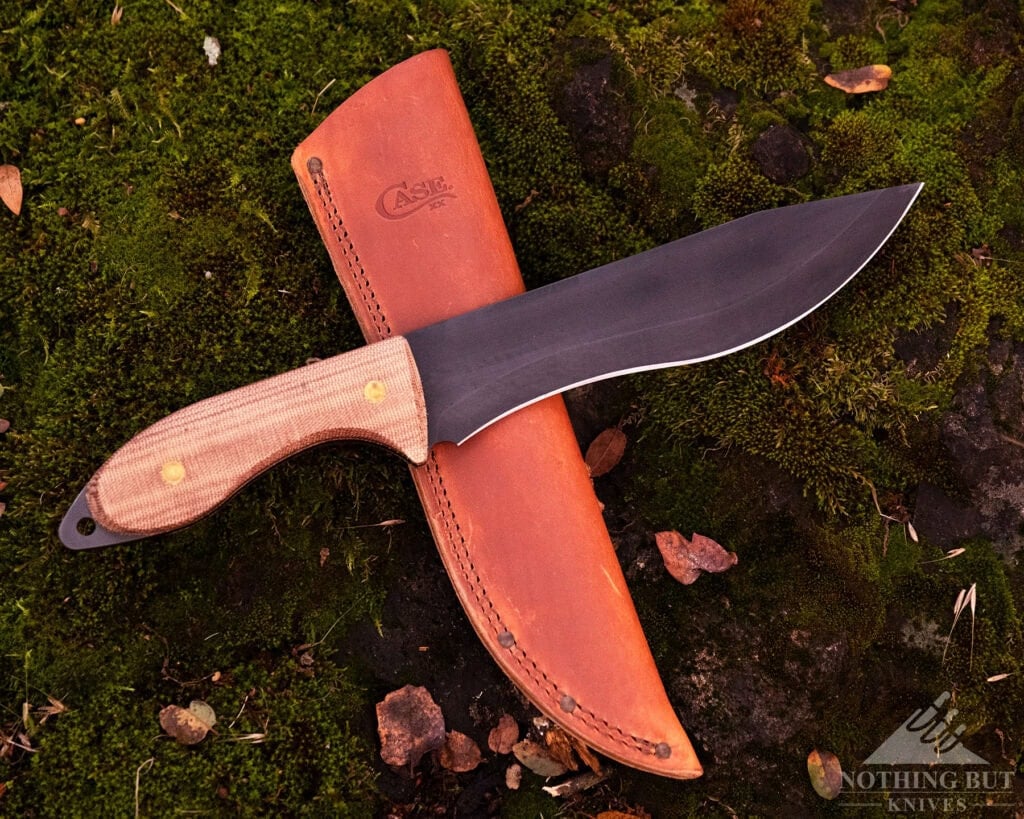 A Few Of Our Favorite Knives With Micarta Handles - Image 5 An overhead view of the Case Knives Laramy Miller Natural Micarta Sasquatch Bowie on top of its leather sheath on a green, moss-covered rock.