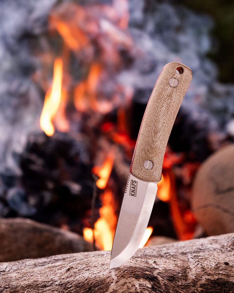 A Few Of Our Favorite Knives With Micarta Handles - Image 6 The Knafs Lulu bushcraft knife with Micarta handle scales and a Magnacut steel blade sticking out of a log next to a campfire in the Sierra Nevada Mountains.