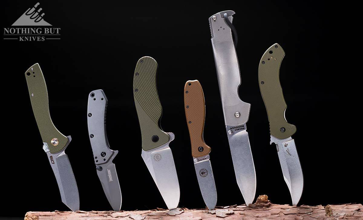 Six of the best hard use pocket knives we have tested sticking out of a piece of wood under moody lighting.