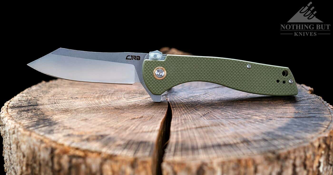 Best Hard Use Pocket Knives Under $50 And $100 - Image 17 The CJRB Kicker folding knife sitting on a tree stump in the open position.