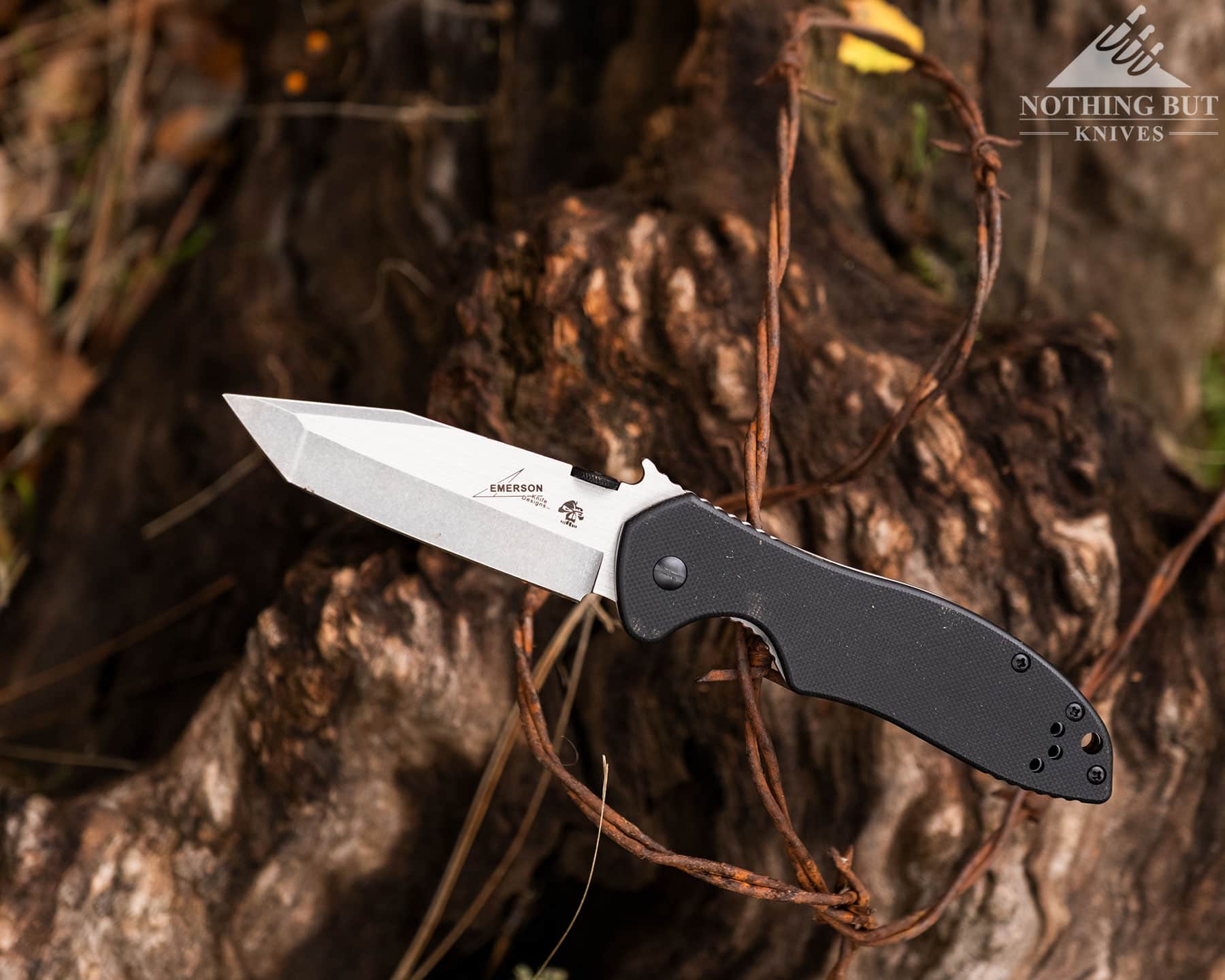 The Best Tactical Knives From Our Favorite Brands - Image 39 An elevated view of the Kershaw Emerson CQC-7K tactical folding knife with its blade in the open position on a section of rusty barbed wire fence above a log.
