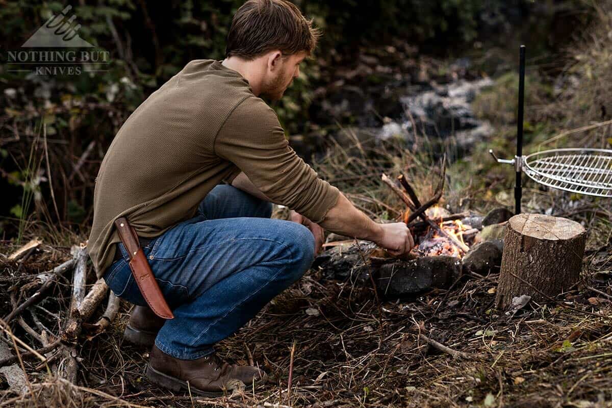 A man tending a camp fire with the Old Hickory butcher knife in a leather sheath on his waist. 