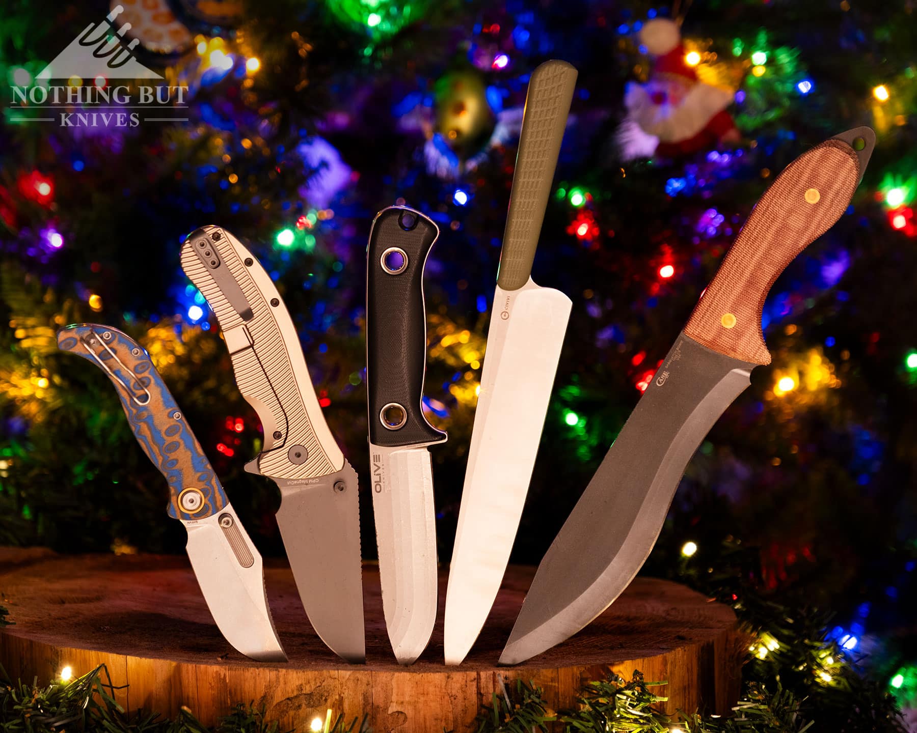 Five knives that would make great gifts shown here on a cedar wood round in front of a decorated Christmas tree.