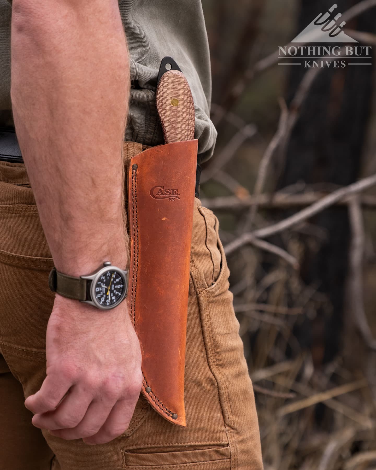 A close-up of the Case Laramie Miller Sasquatch Bowie knife in its brown, leather sheath attached to a belt on a person's waist.