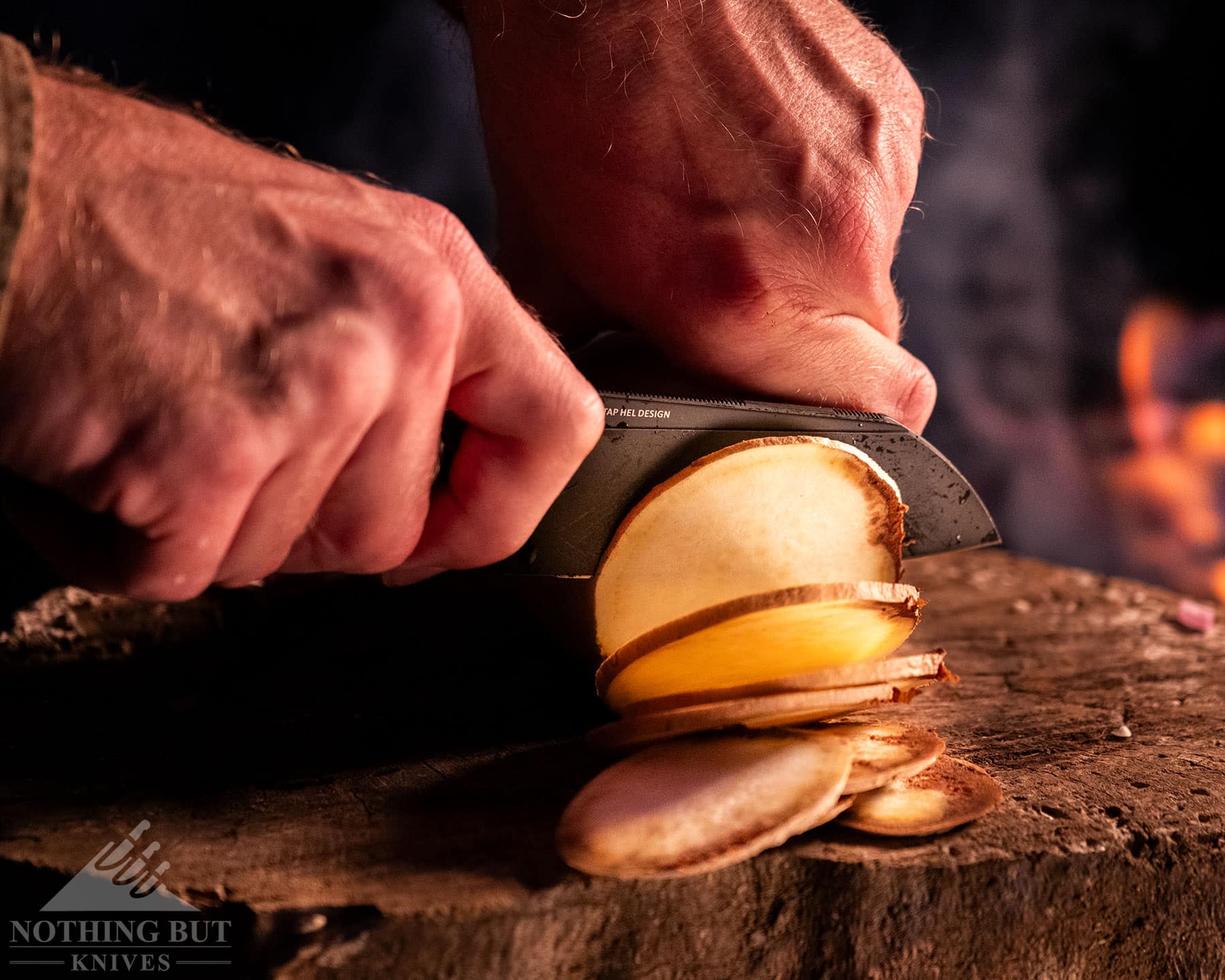 A slightly elevated view of the Civivi Hid folding knife being used to slice a potato next to a campfire.