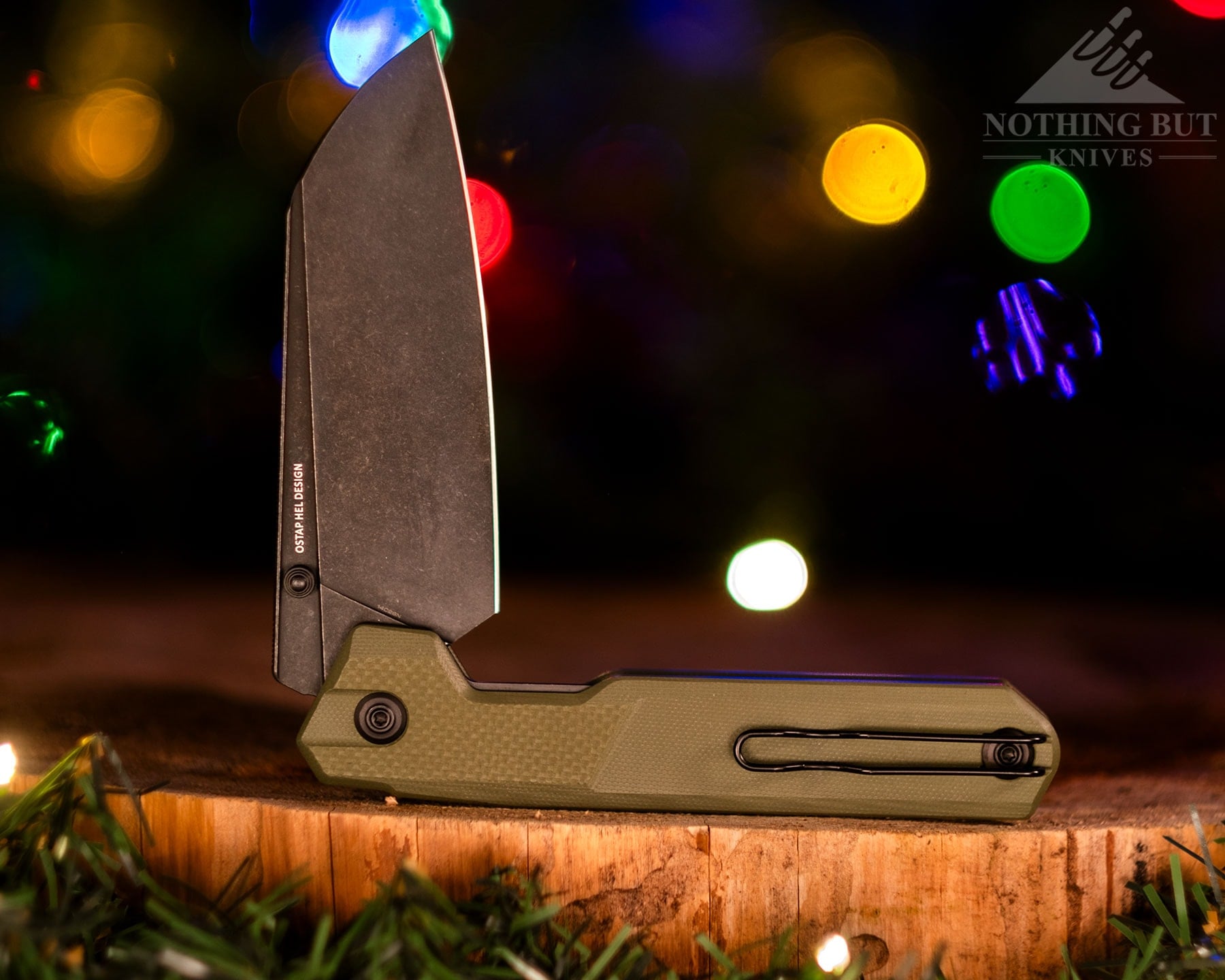 A close-up of the Civivi Hid  camp cooking knife shown here with its blade in the half open position on a piece of wood in front of blurry Christmas lights. 