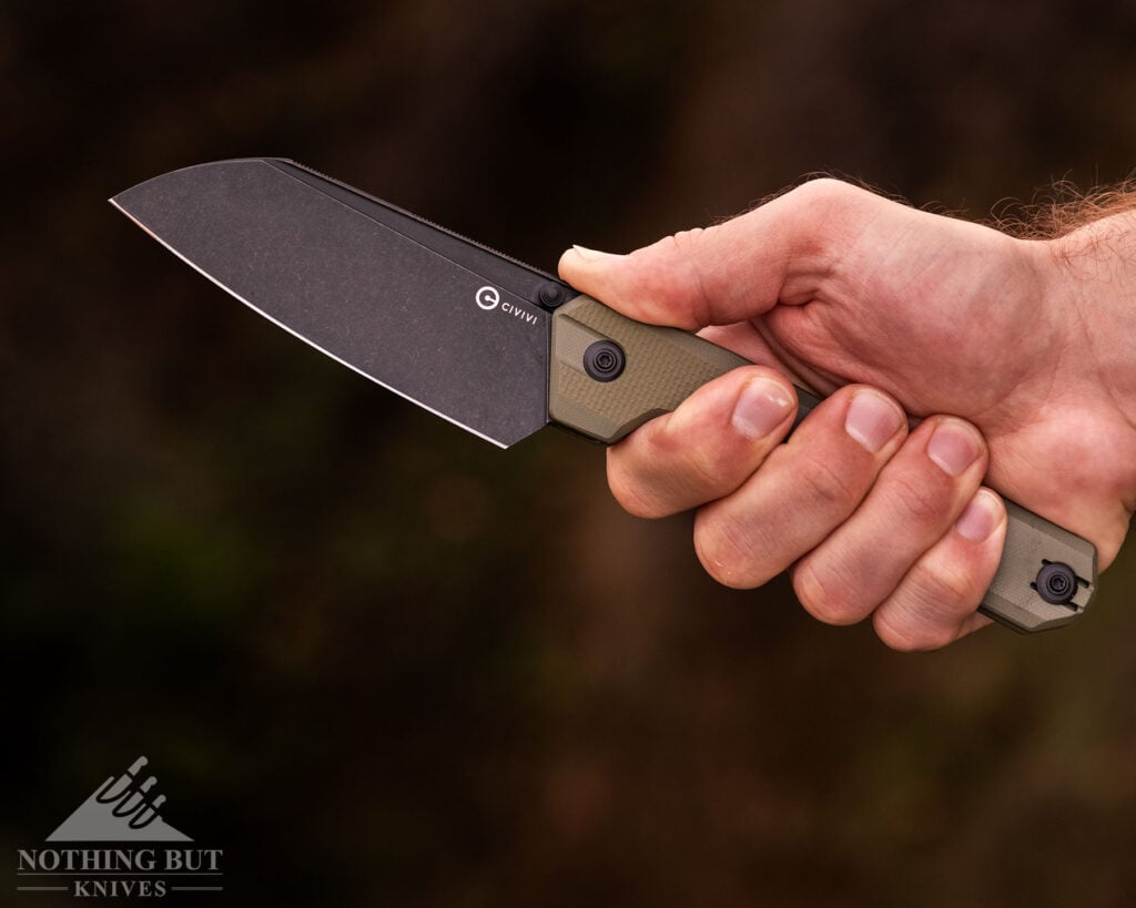 A close-up of the Civivi hid camp knife being held in a person's right hand in front of a blurry, green background. 
