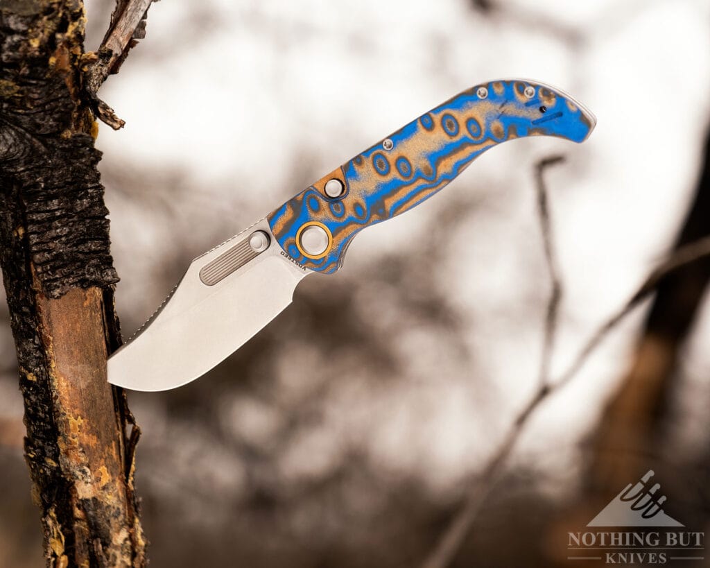 A Bowie styled folding knife from Vosteed shown here with its blade stuck into a fallen tree branch under an overcast sky. 