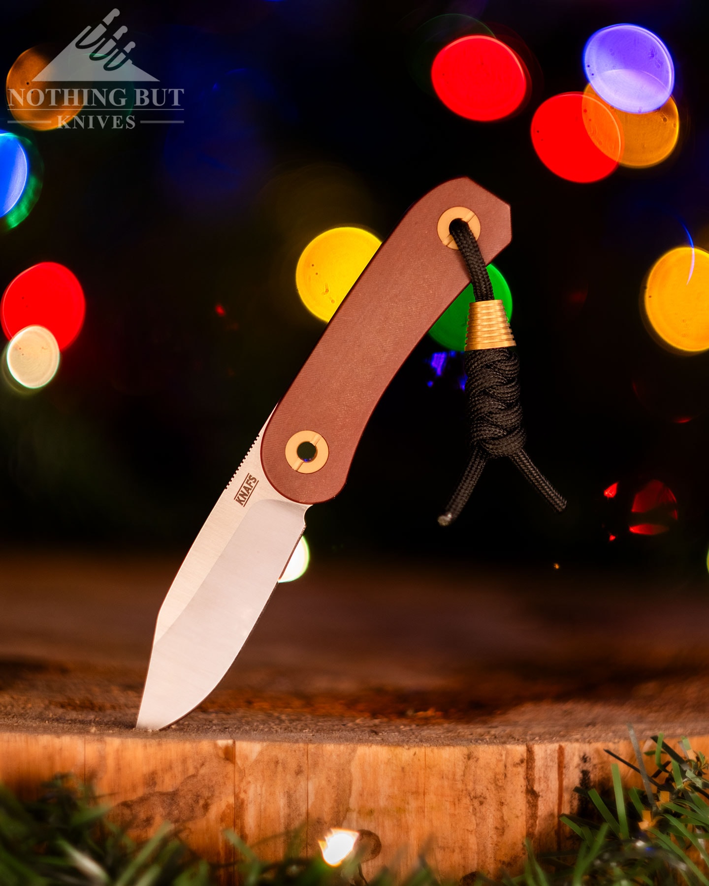 great gift knife from Knafs called the Lander 4 shown here with its blade tip sticking out of a piece of wood in front of blurry holiday lights. 