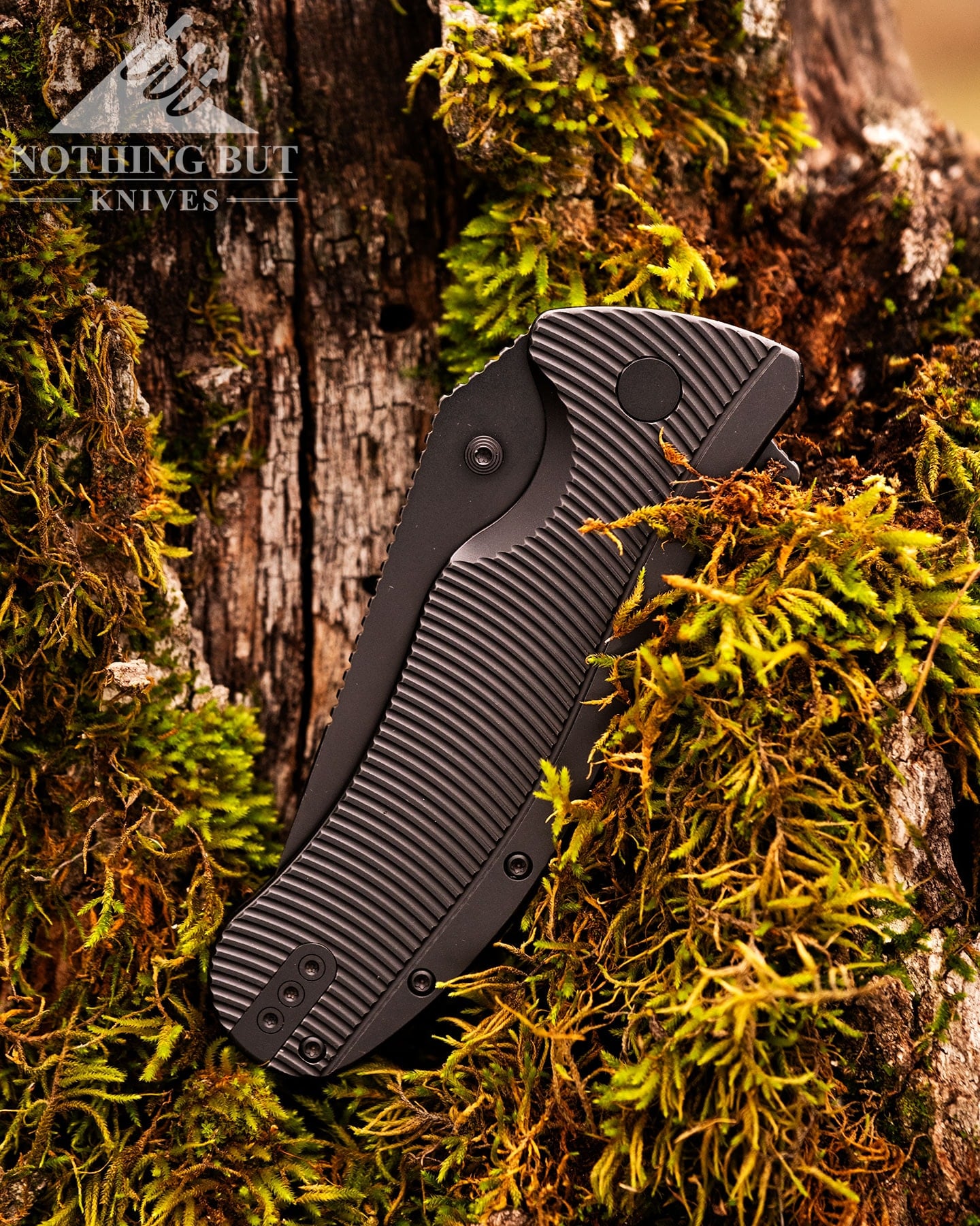 A close-up of a blackout version of the Off-Grid Rhino Elite knife in the closed position balanced in the bark of a moss-covered tree stump. 