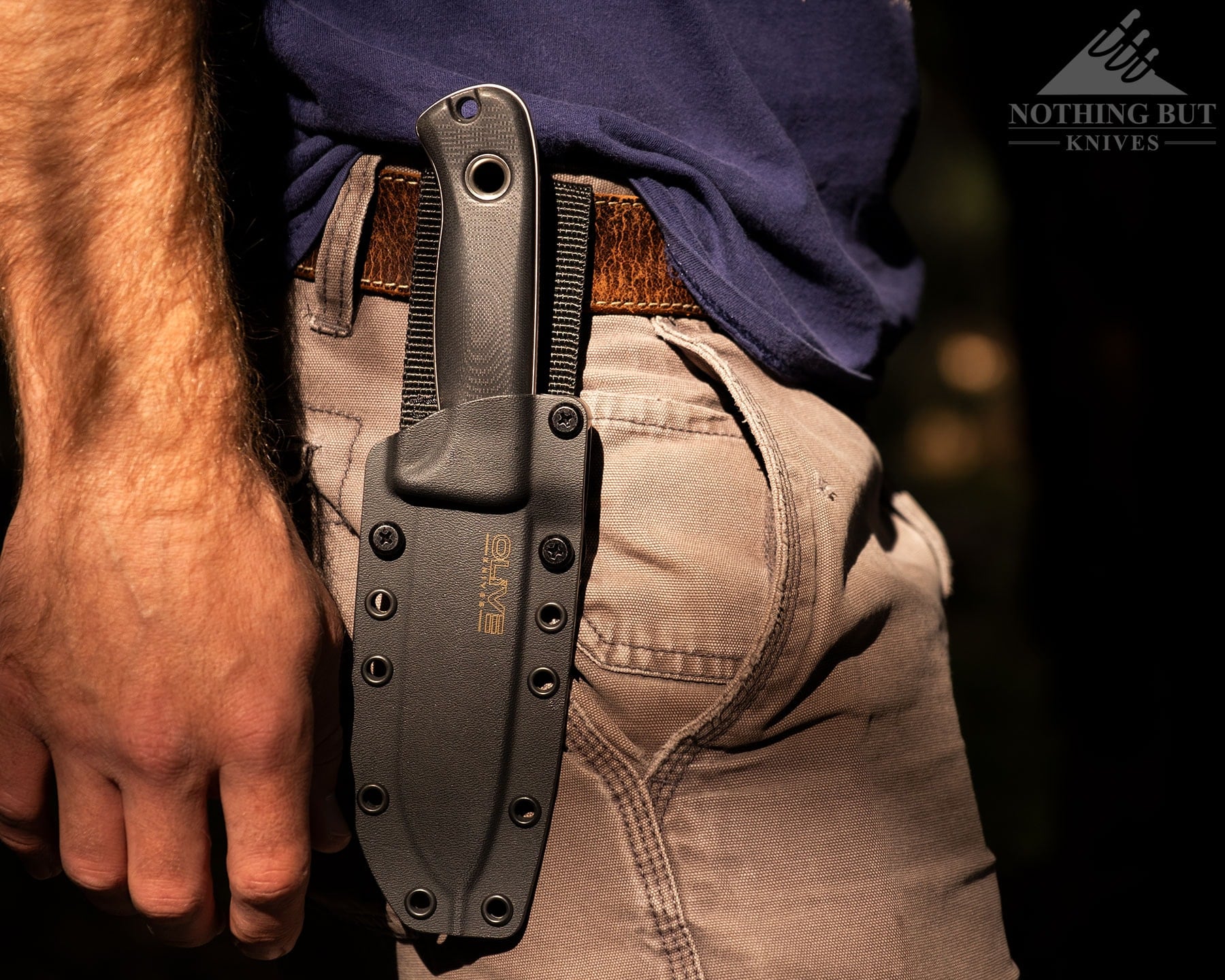 A close-up pf the Olive Arbor fixed blade knife in its kydex sheath attached to a leather belt on a person's waist. 