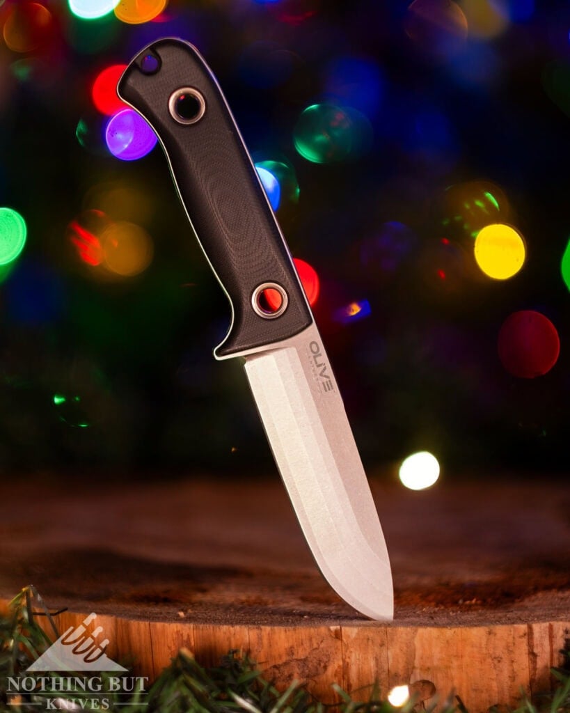 A shallow depth of field image of the Olive Arbor Bushcraft knife sticking out of a cedar round in front of a Christmas tree.