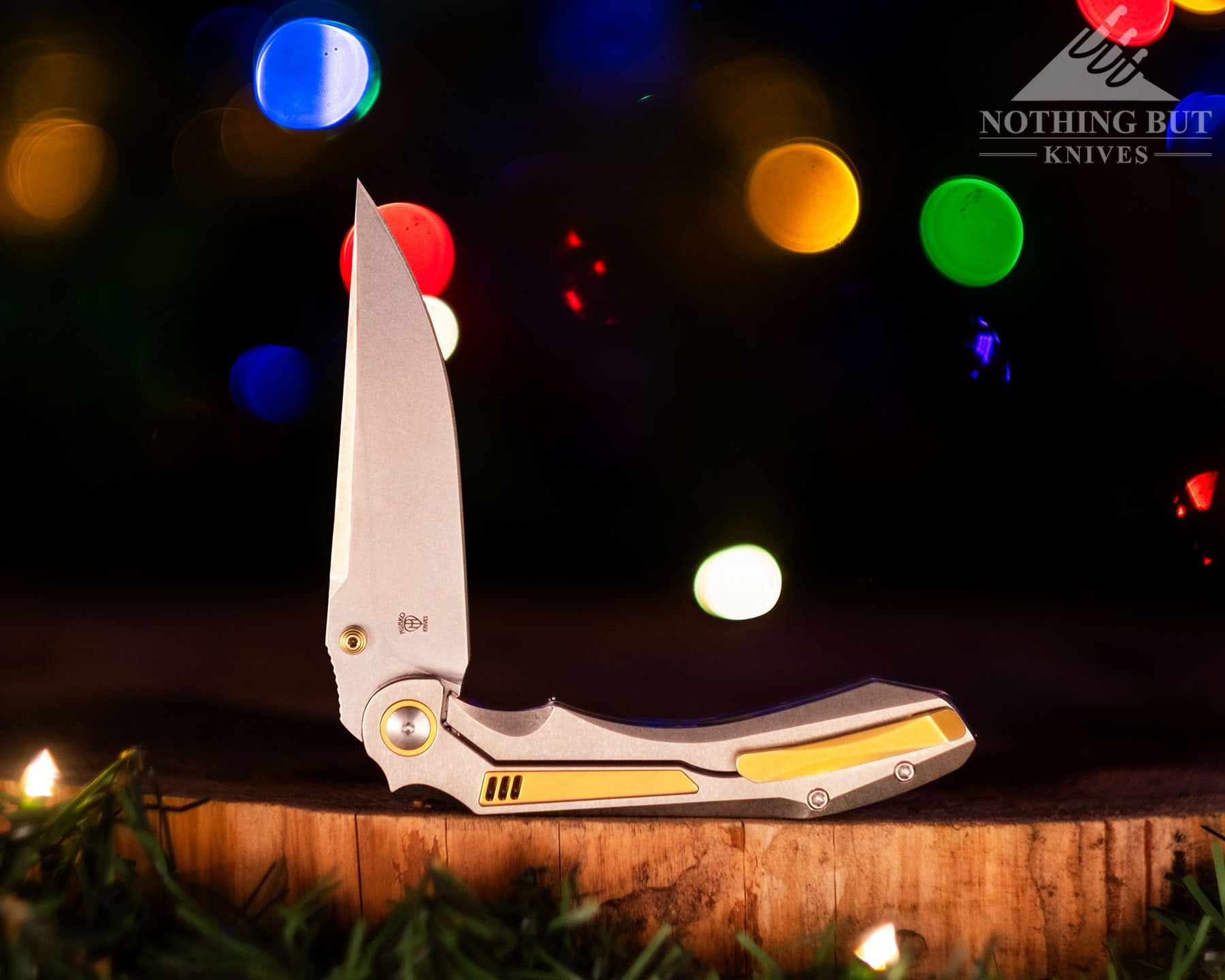 The Orbital Knives Fornax pocket knife shown here with its blade in the half open position on a cedar round in front of glowing holiday lights.