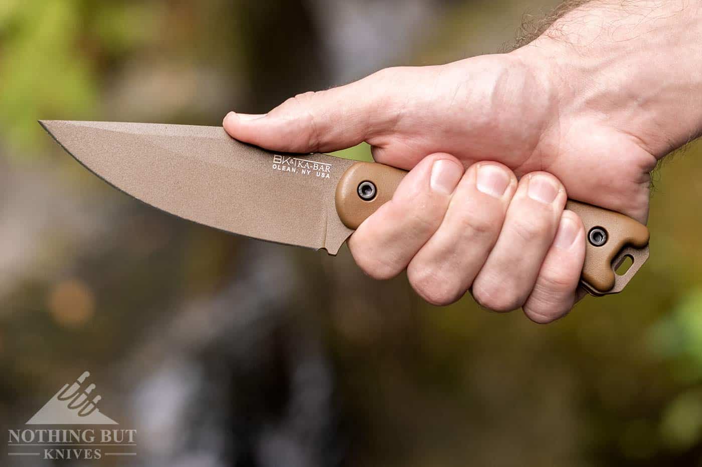 A man's right hand gripping a Ka-Bar survival knife while stablizing it with his thumb pressed firmly against the knife's spine.