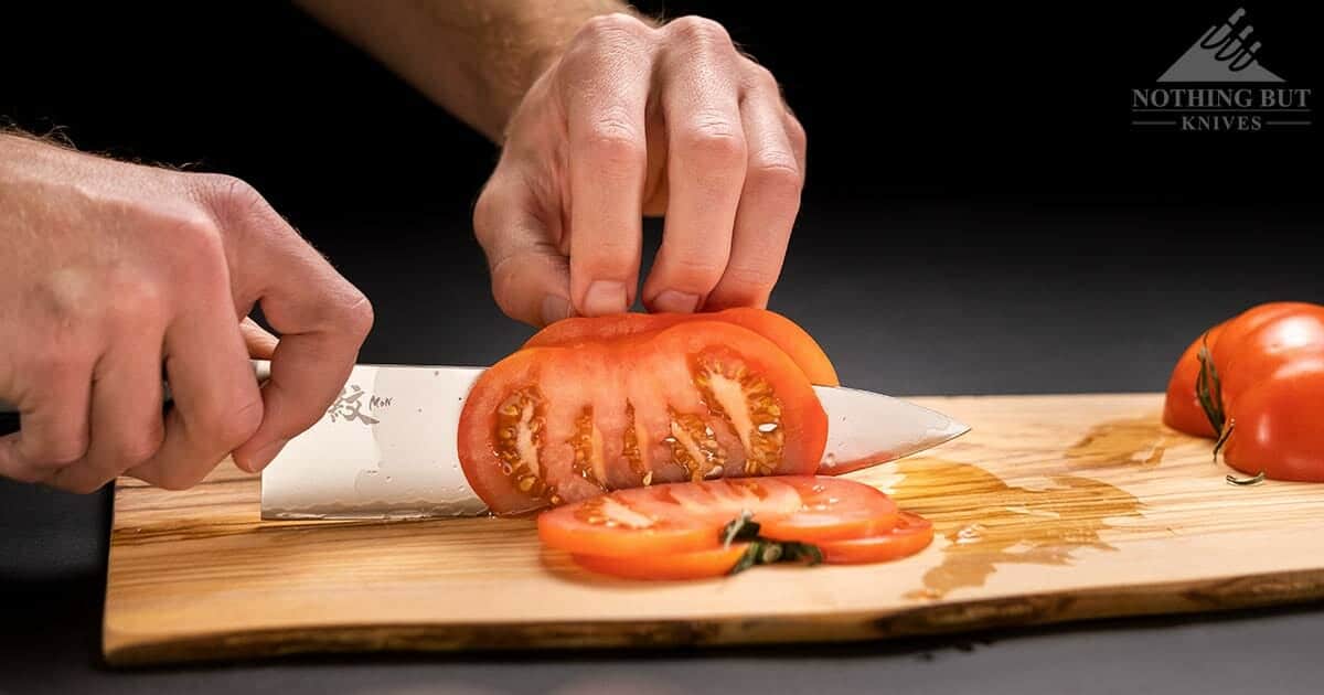 A Yaxell Mon chef knife slicing a tomato. on a wooden cutting board on top of a black background. 