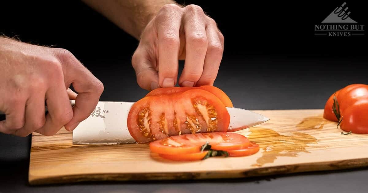 Yaxell Knives And Knife Set Buyers Guide - Image 4 A Yaxell Mon chef knife slicing a tomato. on a wooden cutting board on top of a black background.