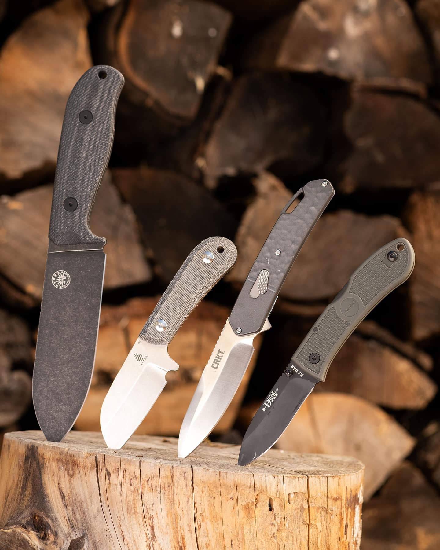 Two folding knives and two fixed knives made of D2 steel lined up on a stump in front of a pile of firewood