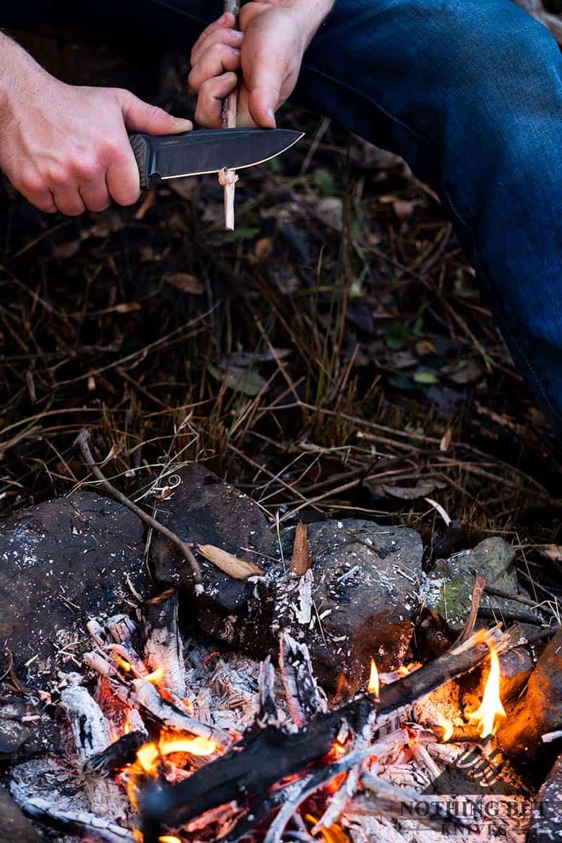 A man's hands carving a stick next to a campfire. 