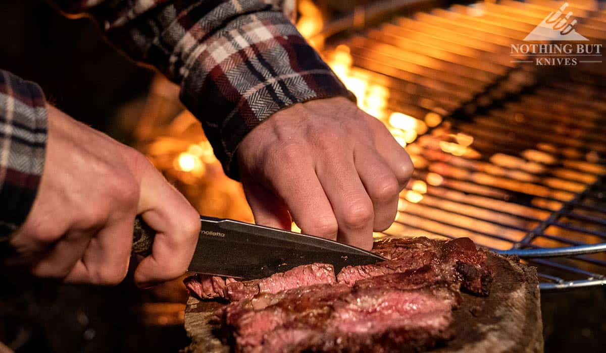 A man's hands using the Lionsteel T5 to slice up a piece of steak next to a campfire. 