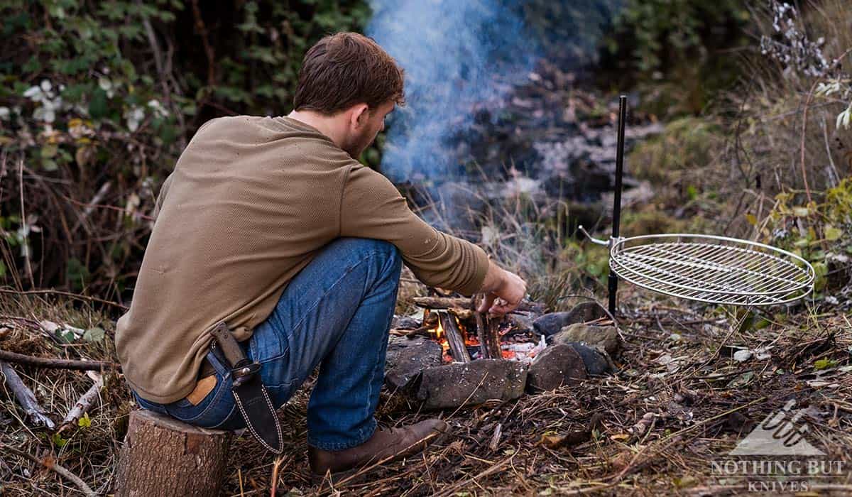 A man tending a campfire with a survival knife in a sheath attached to his belt. 