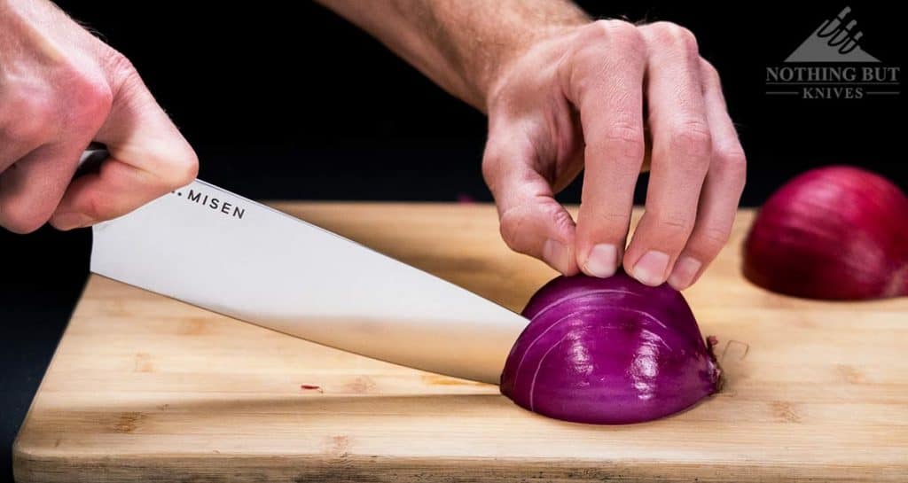 The Misen chef knife slicing through a red onion on a wood cutting board.