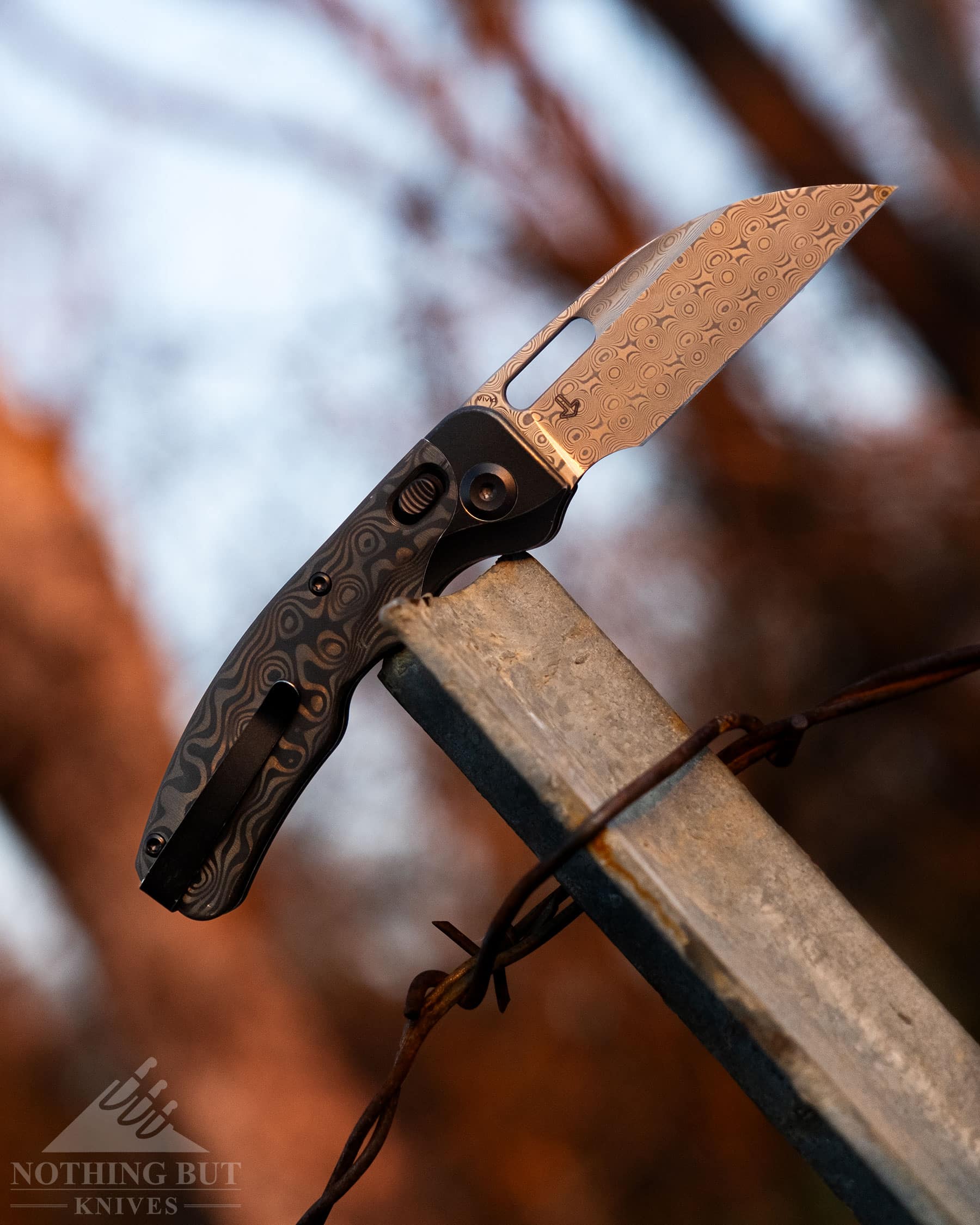 The Kansept Vivid folding knife with Damascus steel and carbon fiber handle scales balanced on a metal fence post in the forest. 