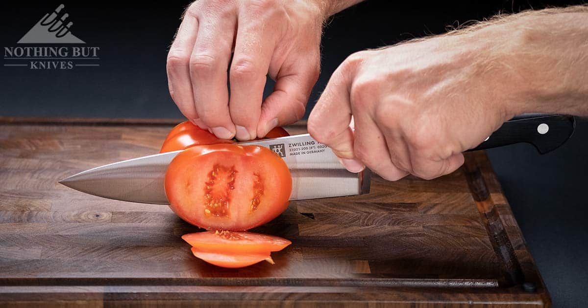 The Zwilling Professional S chef knife slicing through a tomato on an end grain cutting board in front of a dark background. 